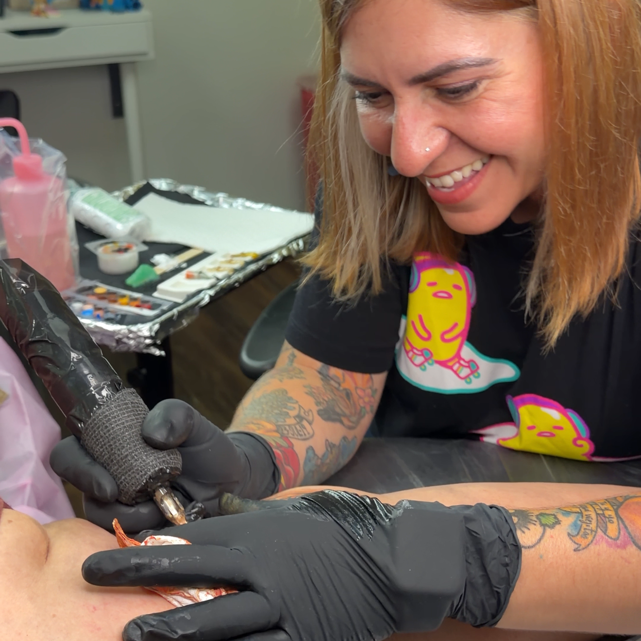 A tattoo artist wearing black gloves and a colorful graphic t-shirt works on a client's shoulder while a ring light illuminates the tattooing area.