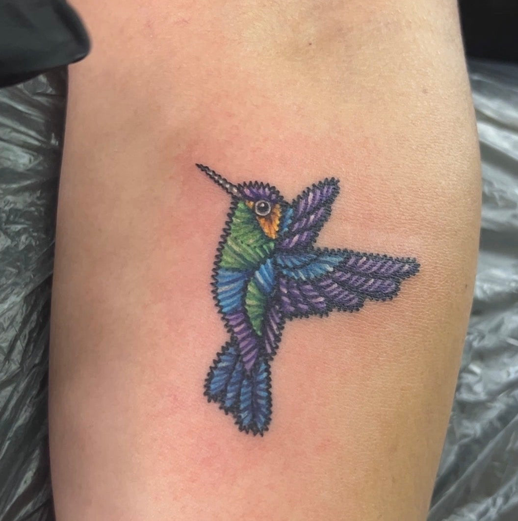 A colorful tattoo of a hummingbird on a person's forearm, featuring purple wings, a green body, and a blue tail, with black plastic sheeting visible in the background.