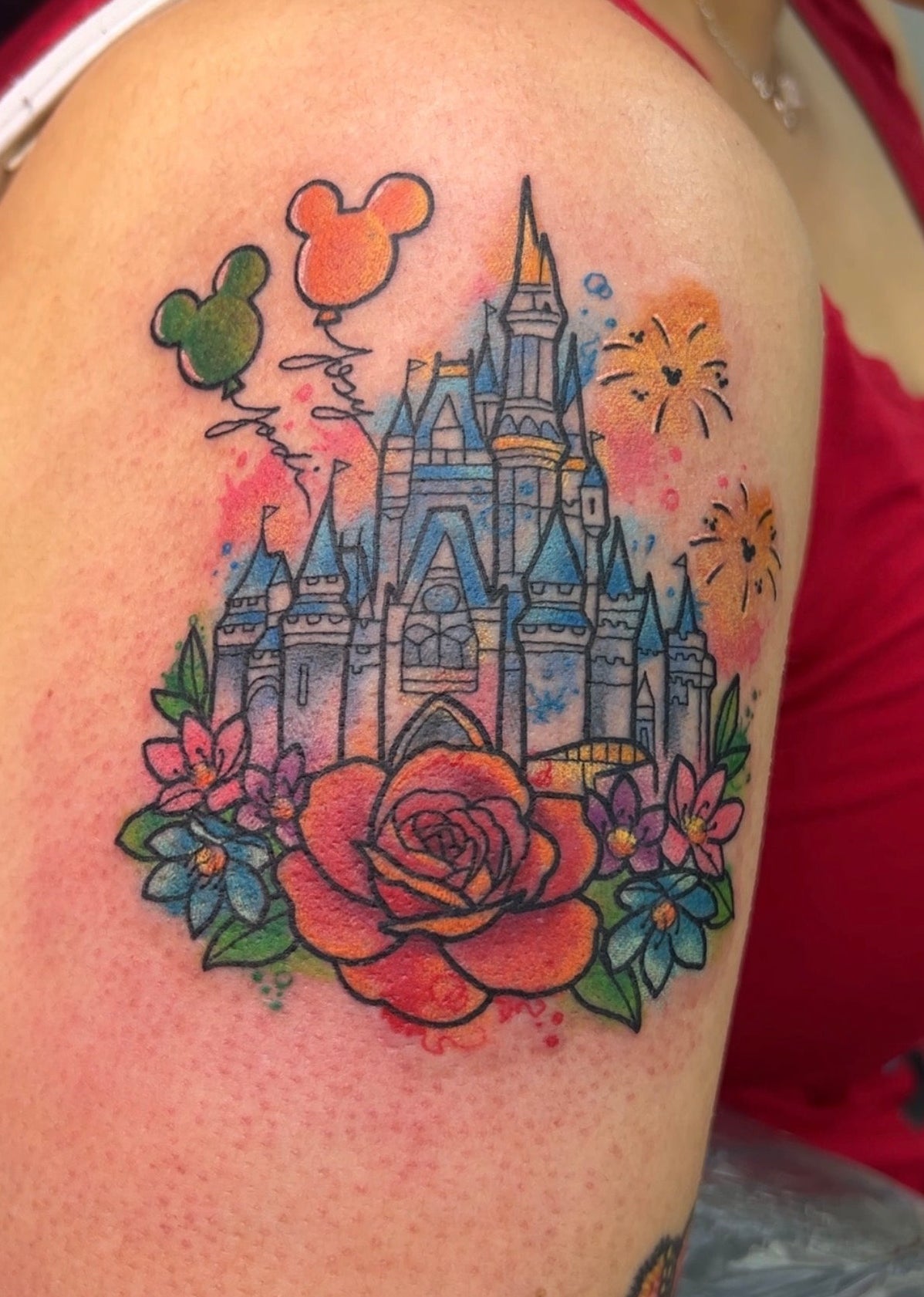 A colorful tattoo on a person's upper arm featuring a castle with Mickey Mouse-shaped balloons, watercolor-style splashes, and flowers including a large red rose.