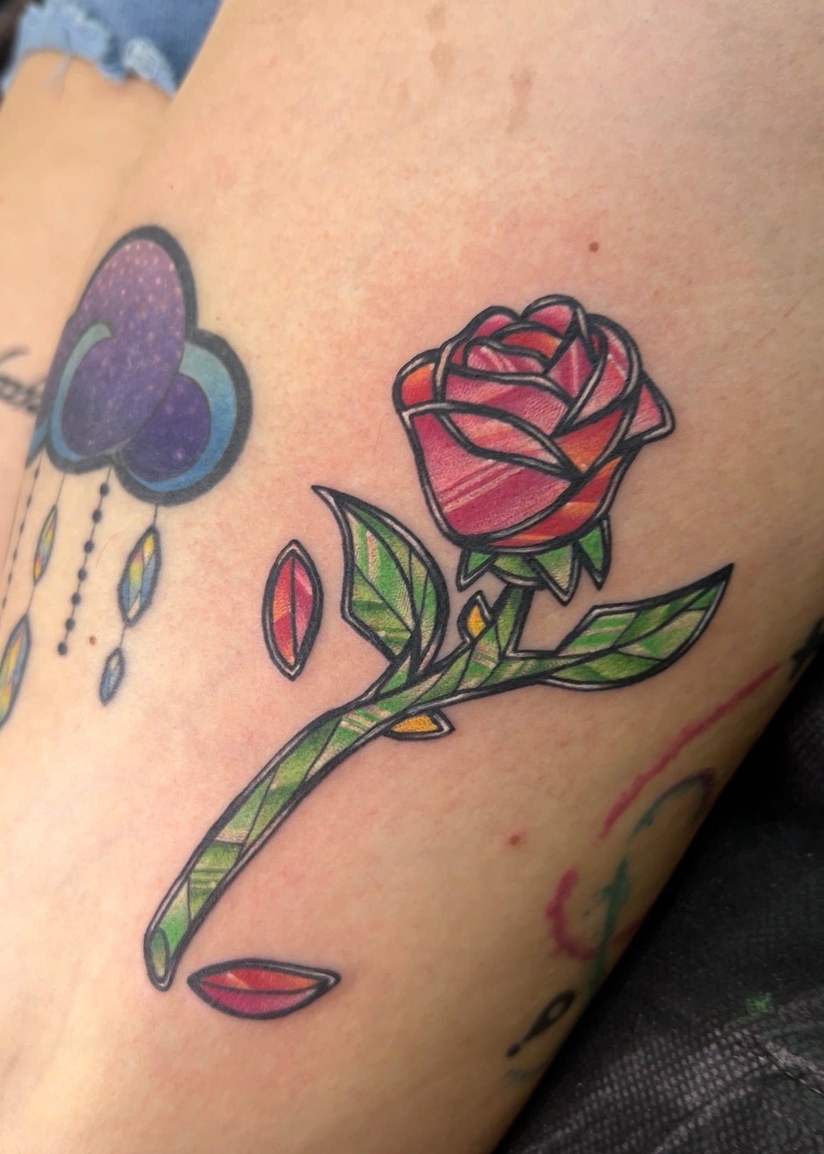A close-up of a tattoo on skin showing a stylized rose with green leaves and pink petals, alongside partial other tattoos including a purple cloud.