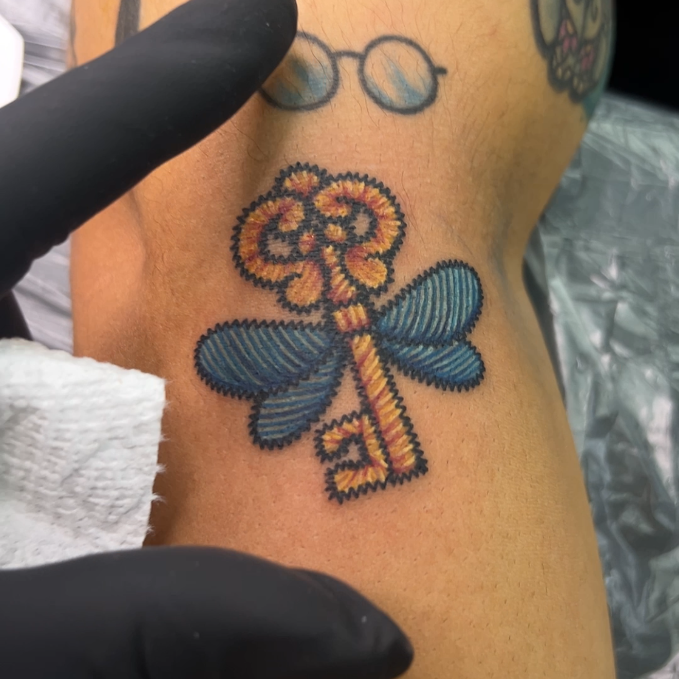 A close-up of a tattoo on a person's forearm featuring a stylized key with blue wings and a gold center, shown with a black-gloved hand pointing at it.