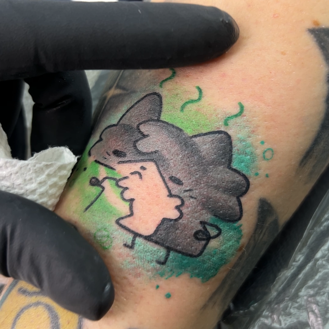 A close-up of a freshly tattooed arm featuring a cartoon dog character with a pink bow and a green watercolor splash background. A black-gloved hand is pointing at the tattoo, with other tattoos and a black plastic sheet visible in the background.