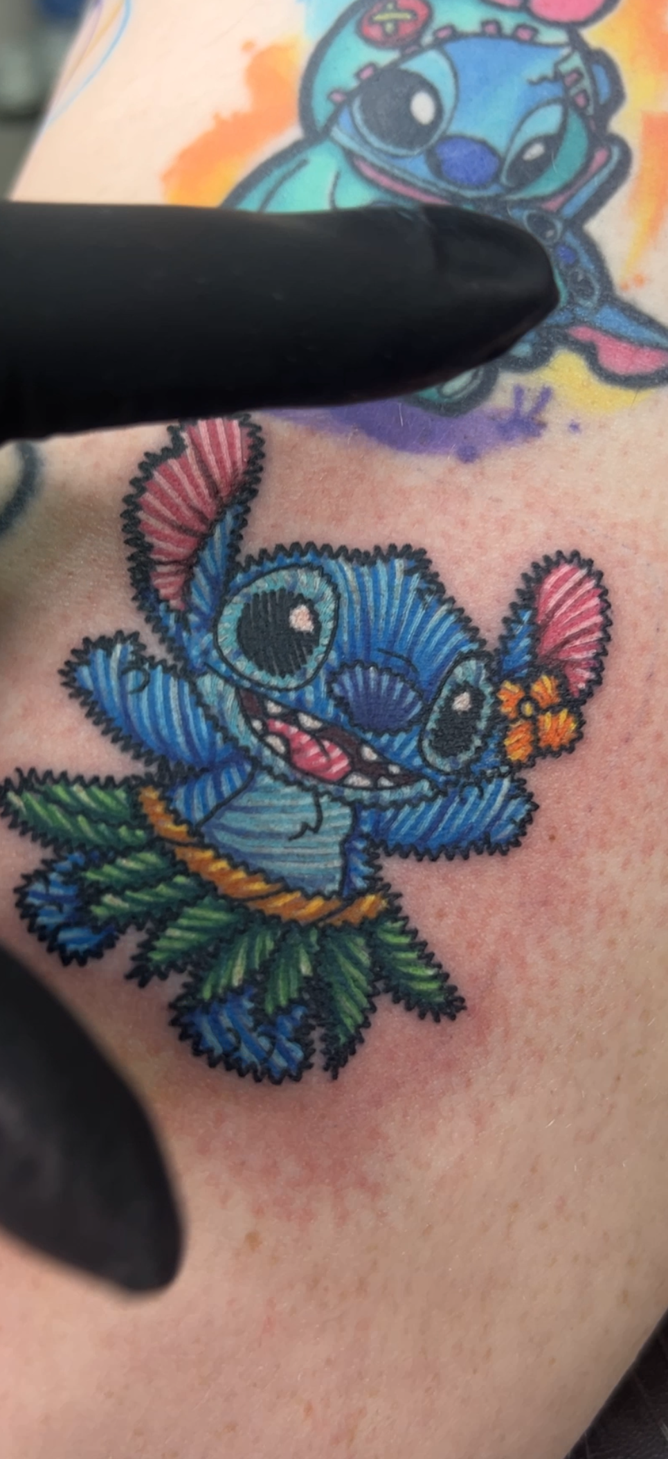 A close-up of a colorful tattoo on skin, showing a cartoon character design being pointed at by a black gloved finger. The tattoo features a blue character with pink ears, a green hula-style skirt, and small orange flower accents.