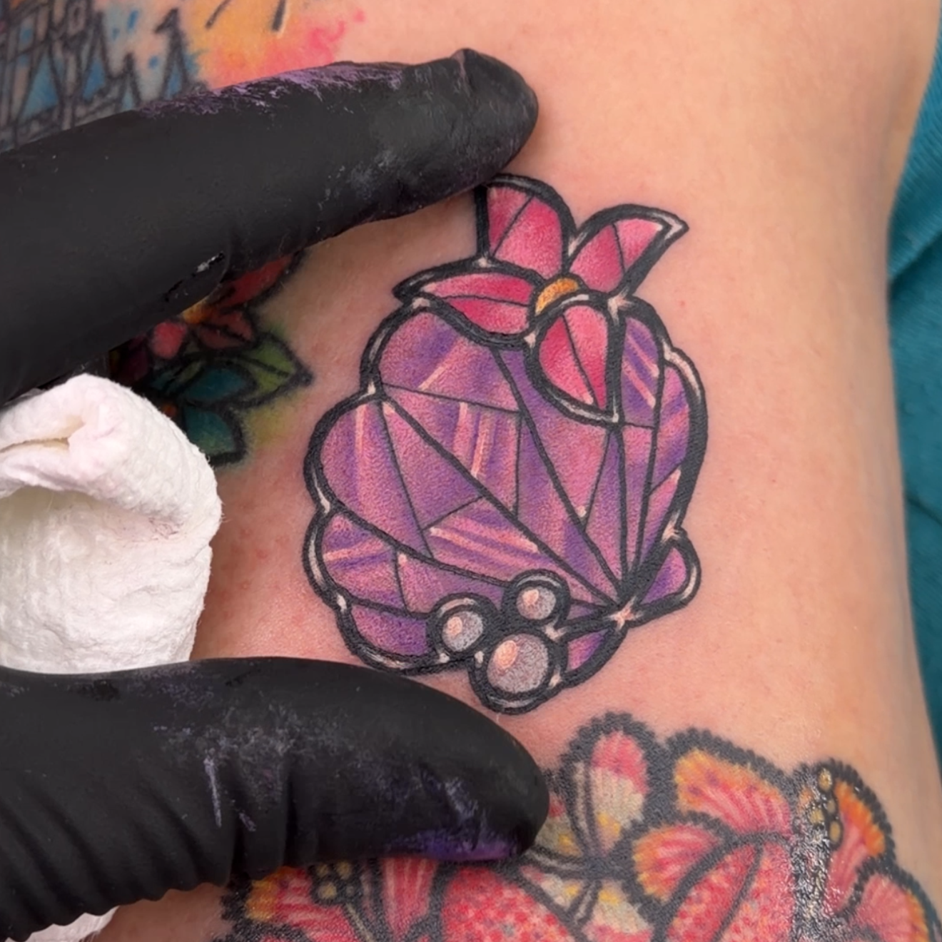 A close-up of a freshly tattooed purple and pink butterfly on a person's upper arm, with a black gloved hand wiping the tattoo area with a white cloth. Other colorful tattoos are visible on the arm.