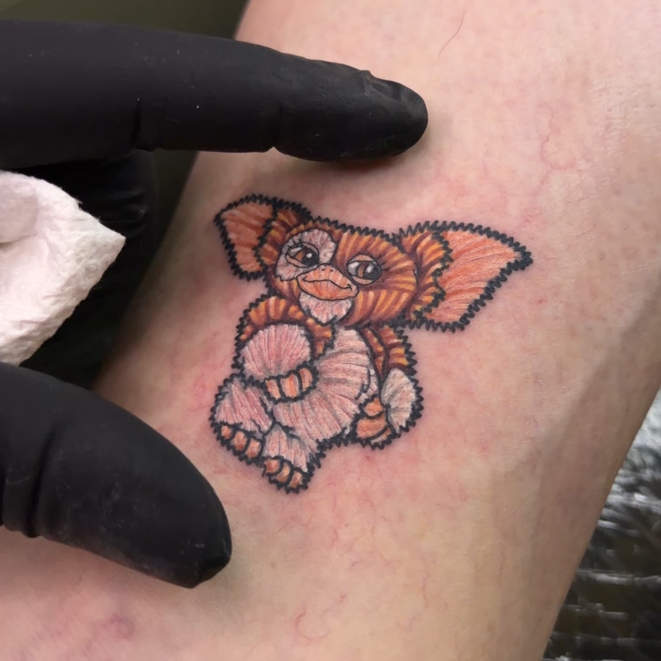 A close-up of a freshly tattooed character on a person's upper arm, with a black gloved hand pointing at the tattoo. The tattoo depicts a small orange, pink, and black creature with large ears.