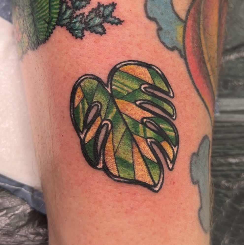 A close-up of a tattooed forearm featuring a green monstera leaf design with yellow and black striping, alongside other colorful tattoos.