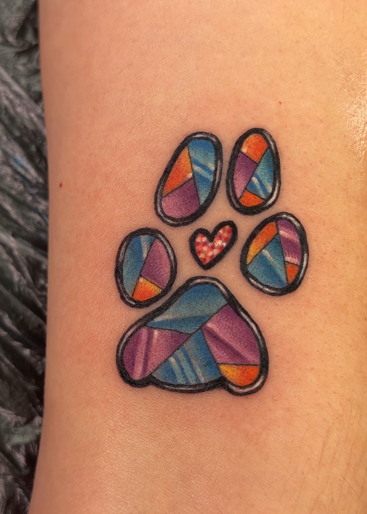 A close-up of a person's skin showing a small paw print tattoo with a colorful mosaic pattern and a small heart in the center.