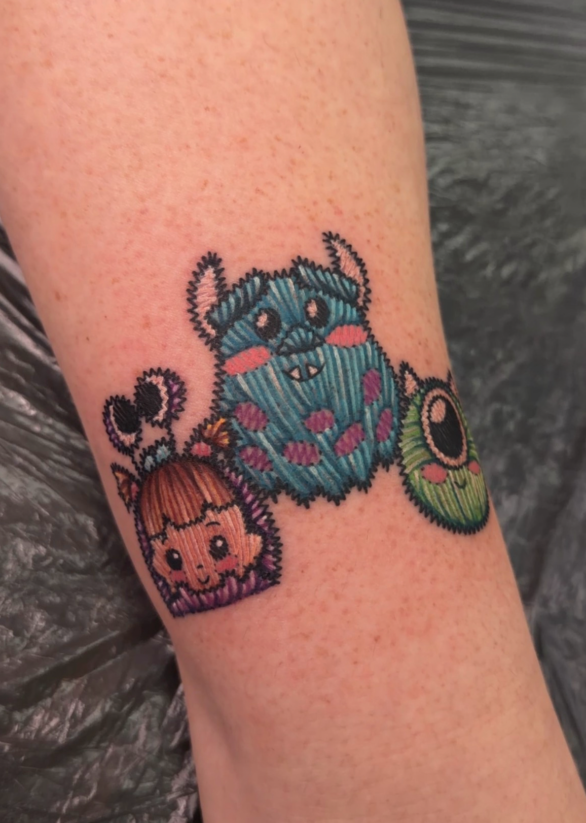 A close-up of a person's forearm featuring a colorful tattoo of three cute, pixel-art style monsters in blue, green, pink, and brown tones.
