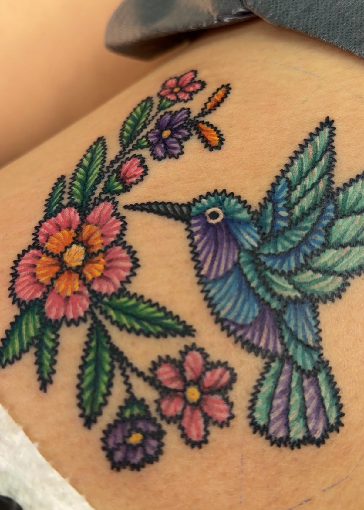 A close-up of a colorful tattoo on skin, featuring a hummingbird in blue, green, and purple tones with pink and purple flowers and green leaves.