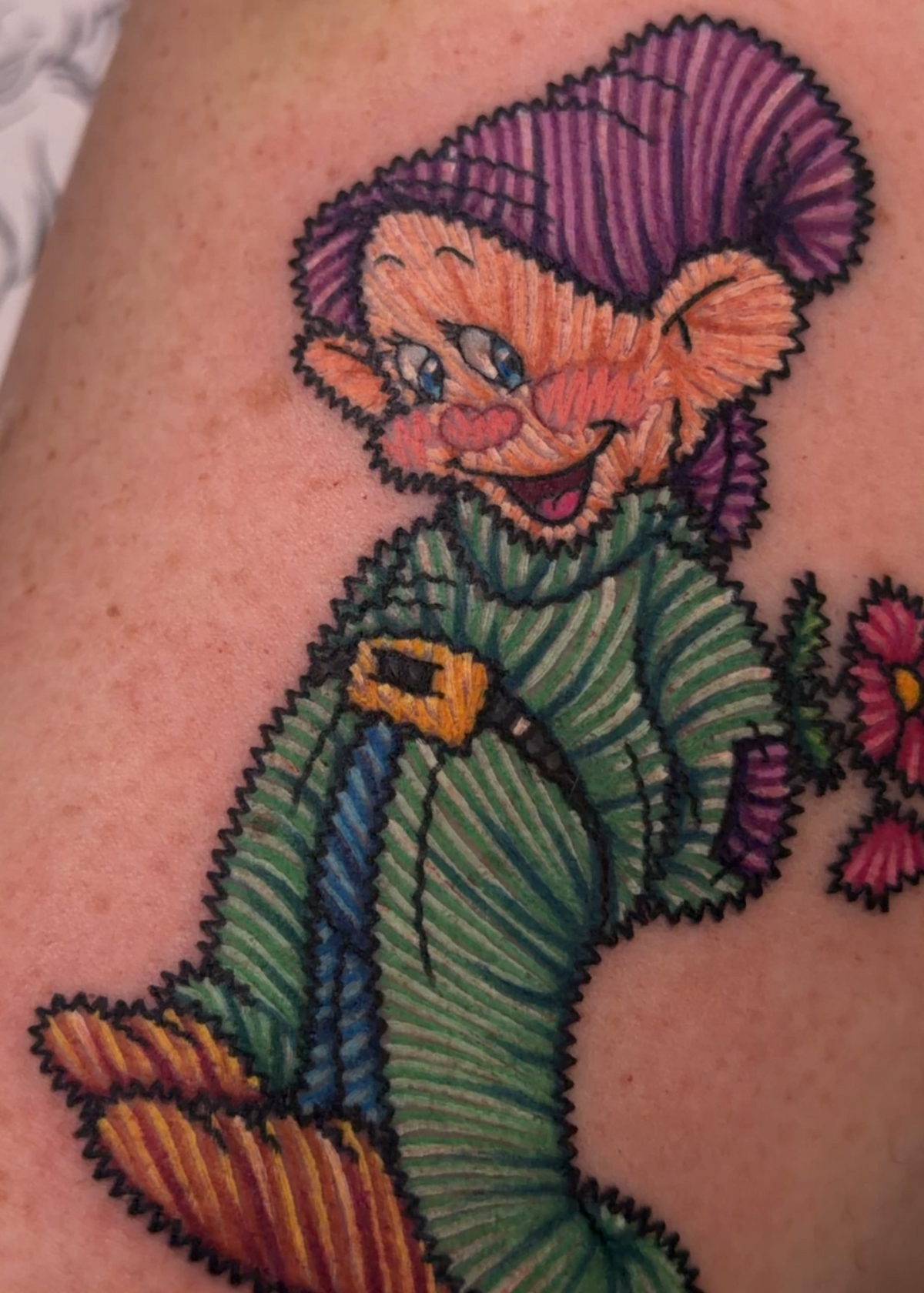 A close-up of a colorful tattoo on skin, depicting a cartoon gnome character with a purple hat, green striped clothing, and orange boots, alongside a small pink flower.