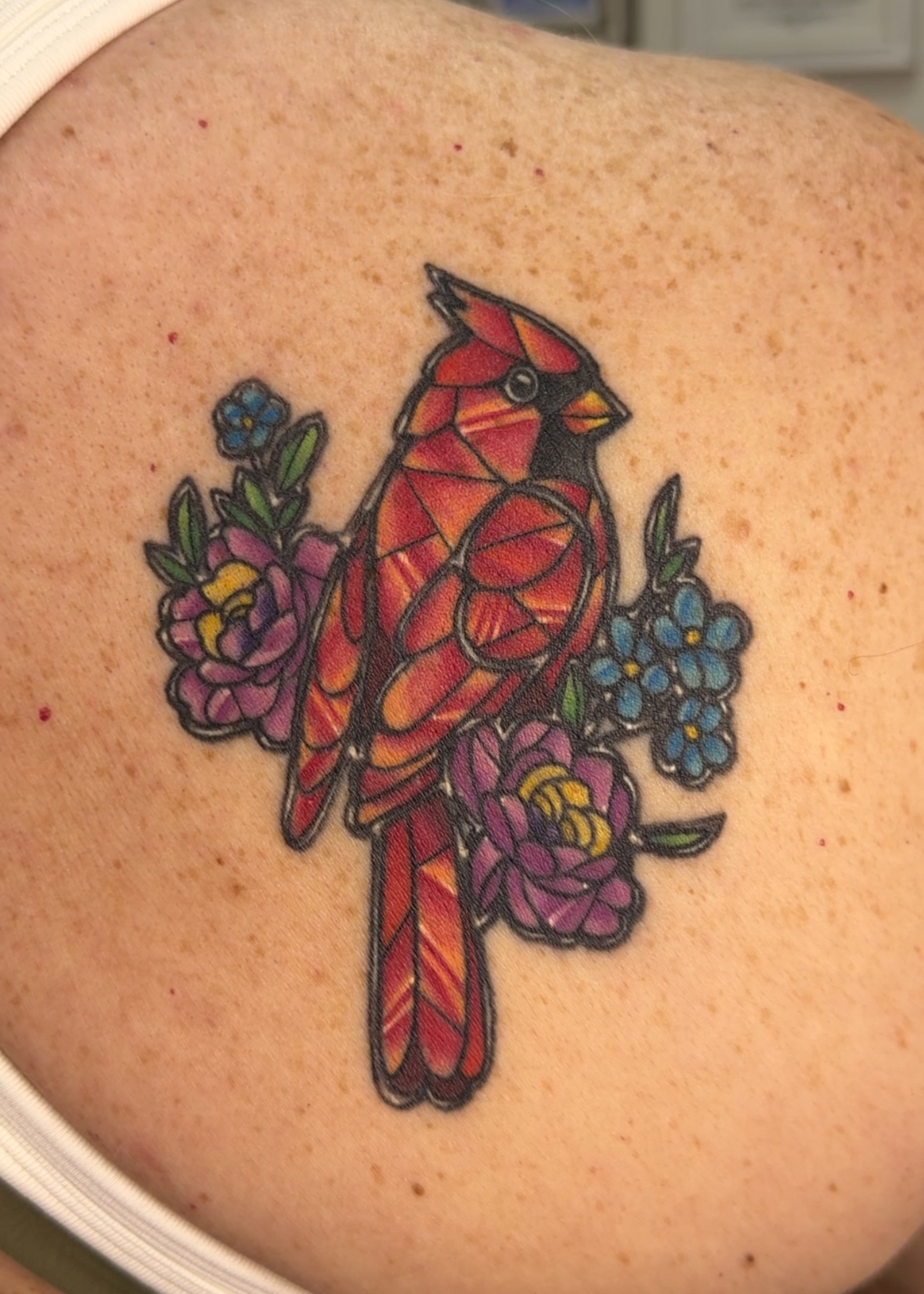 A close-up of a colorful geometric cardinal tattoo on a person's upper back, featuring red, pink, purple, and blue flowers with black outlines.