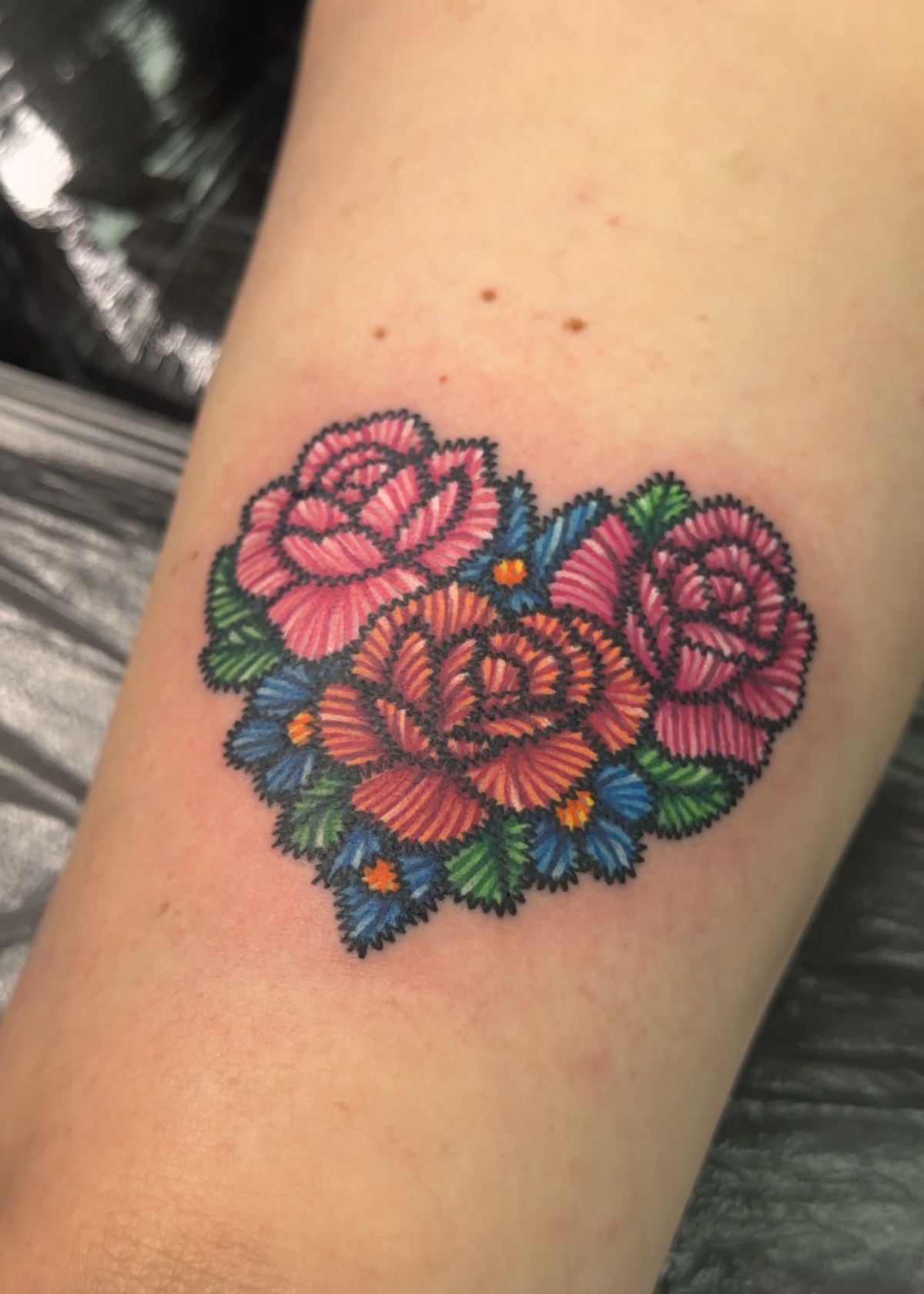 A close-up of a tattoo on a person's forearm, featuring three stylized pink and red roses with blue and green leaves arranged in a heart shape.