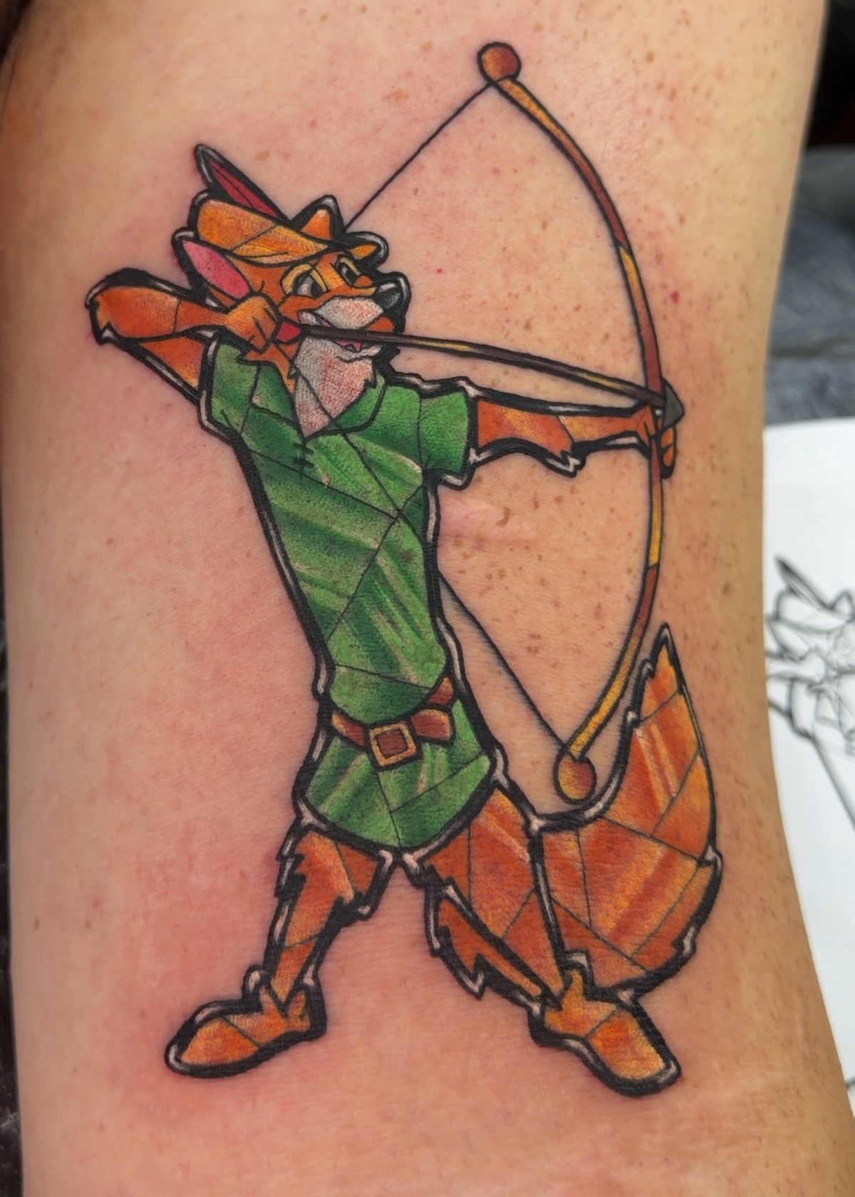 A colorful tattoo of an anthropomorphic fox archer on a person's upper arm, featuring a green tunic, orange tail and boots, and black outlines.