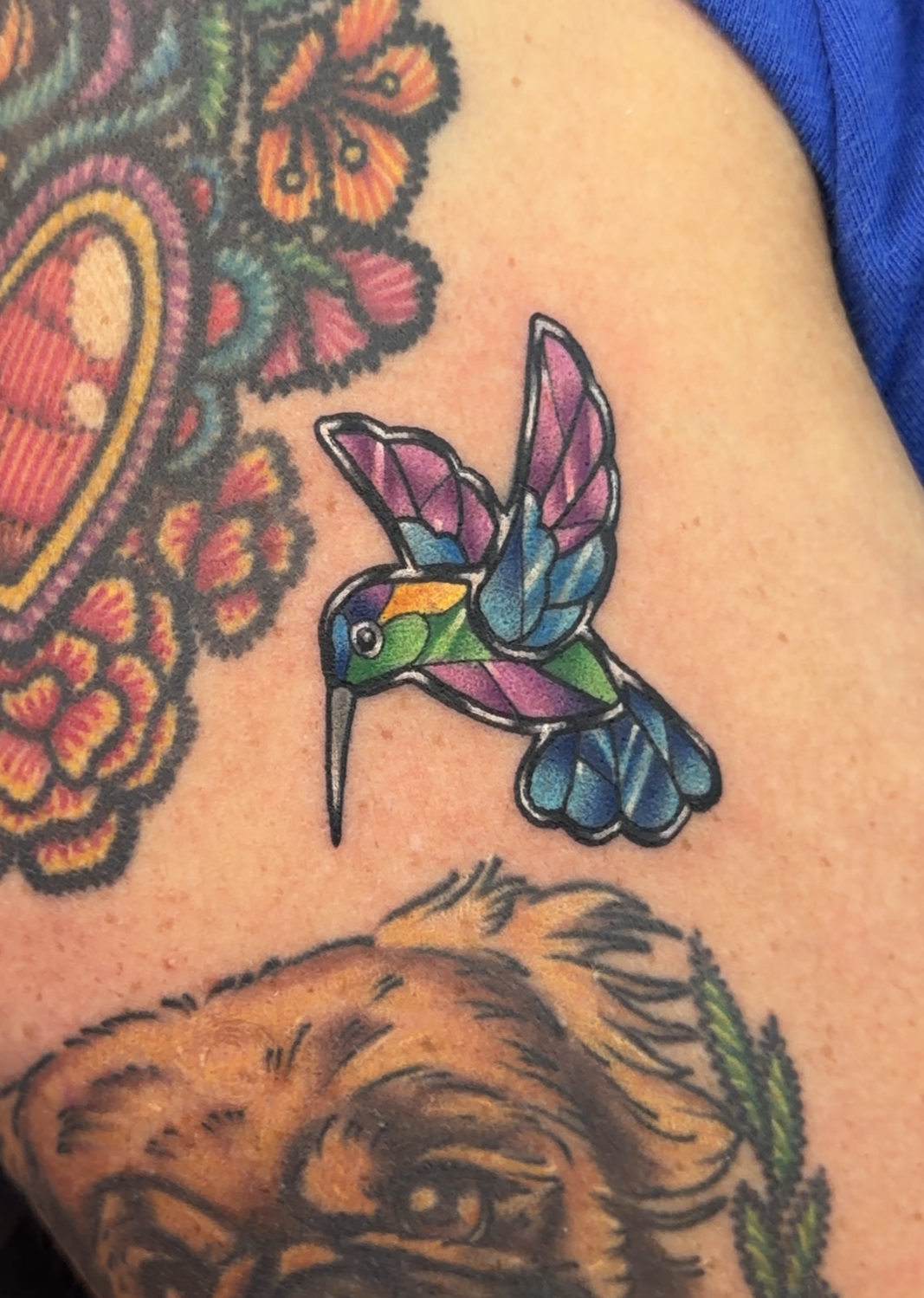 A close-up of a tattooed arm featuring a colorful hummingbird, a heart-shaped flower, a pug portrait, and floral designs, with blue fabric visible at the top.